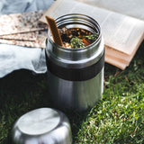 Schwarzer BLACK+BLUM Lunchbecher FOOD FLASK mit orangefarbenem Deckel auf grünem Stoff.