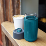 BLACK+BLUM Kaffeebecher TO-GO Glas, blau, offengelegt mit Kaffee, ideal für unterwegs, vor Meerhintergrund.