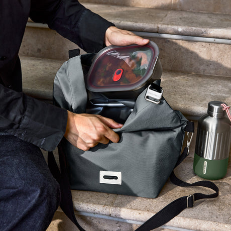 Schwarzer isolierter Lunchbag von BLACK+BLUM mit 10-Liter Fassungsvermögen, sitzend auf Treppen, vertikale Ansicht.